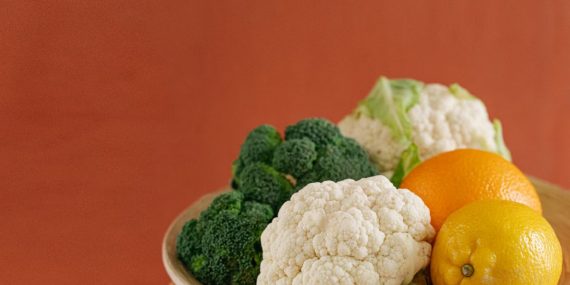Close-up of fresh broccoli, cauliflower, lemon, and orange on a wooden plate with an earthy background.