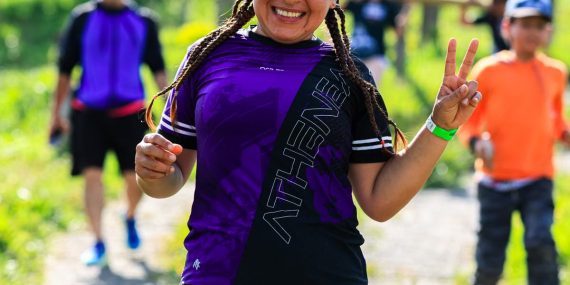 A cheerful female runner flashes a peace sign during an outdoor run with others in a sunny, green setting.
